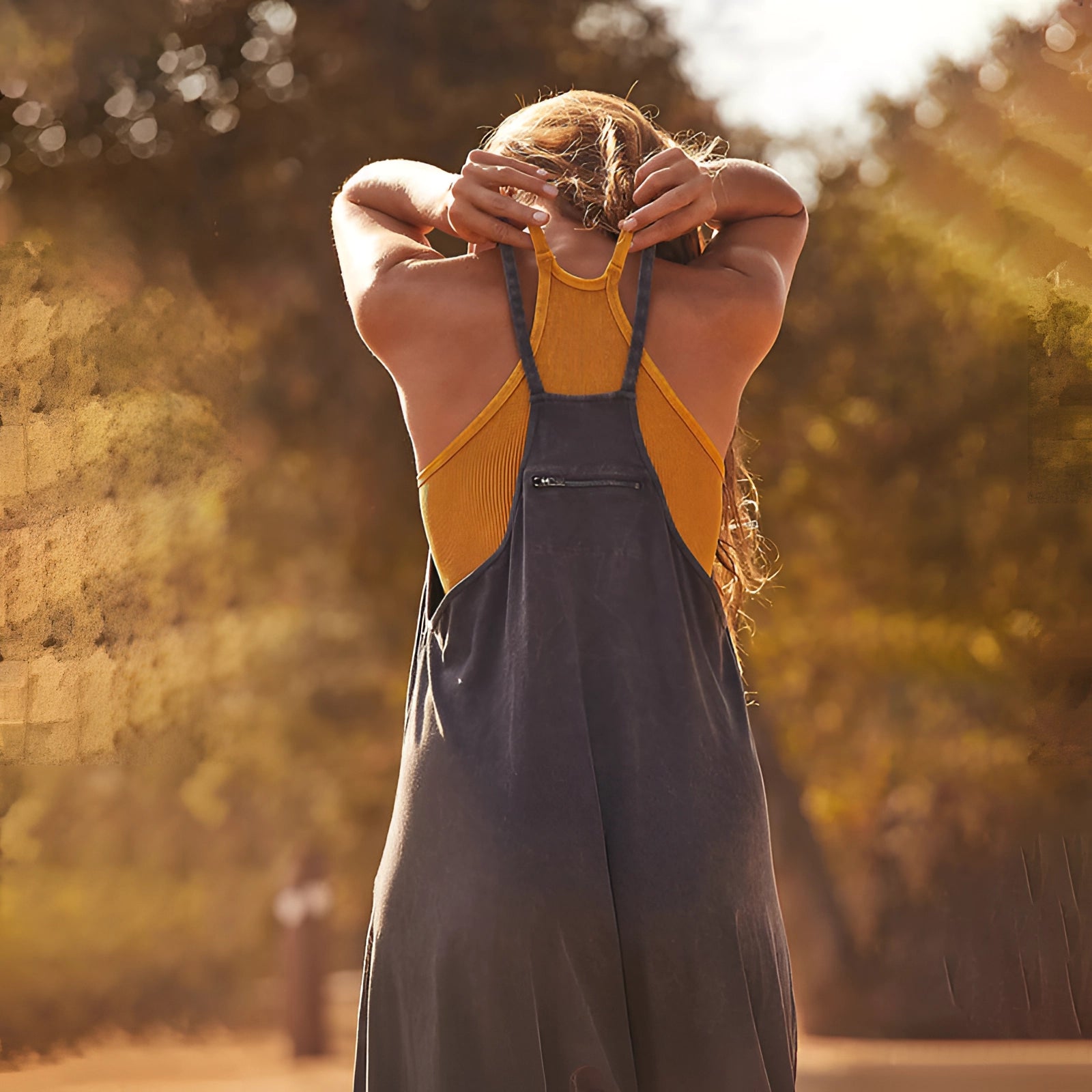 Woman wearing a sleeveless linen jumpsuit, standing in a grassy field under sunlight, smiling and looking happy, surrounded by nature.